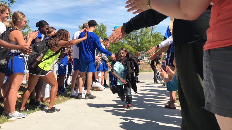 A child, wearing a backpack and holding a sweatshirt,  walks through a crowd of people lined up giving them high-fives