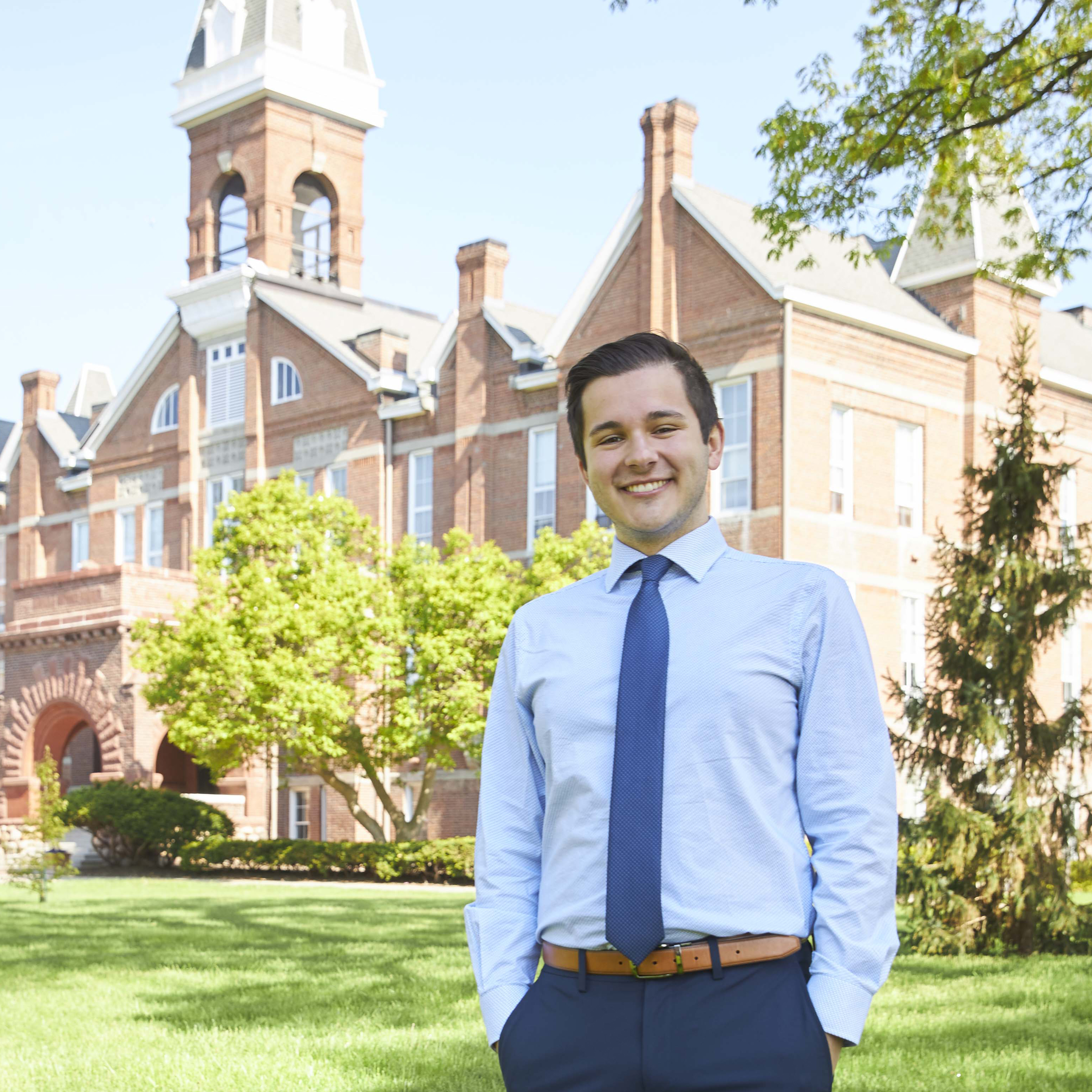 A Drake student in a dress shirt and tie, hands in pockets, stands near a brick building on a sunny day