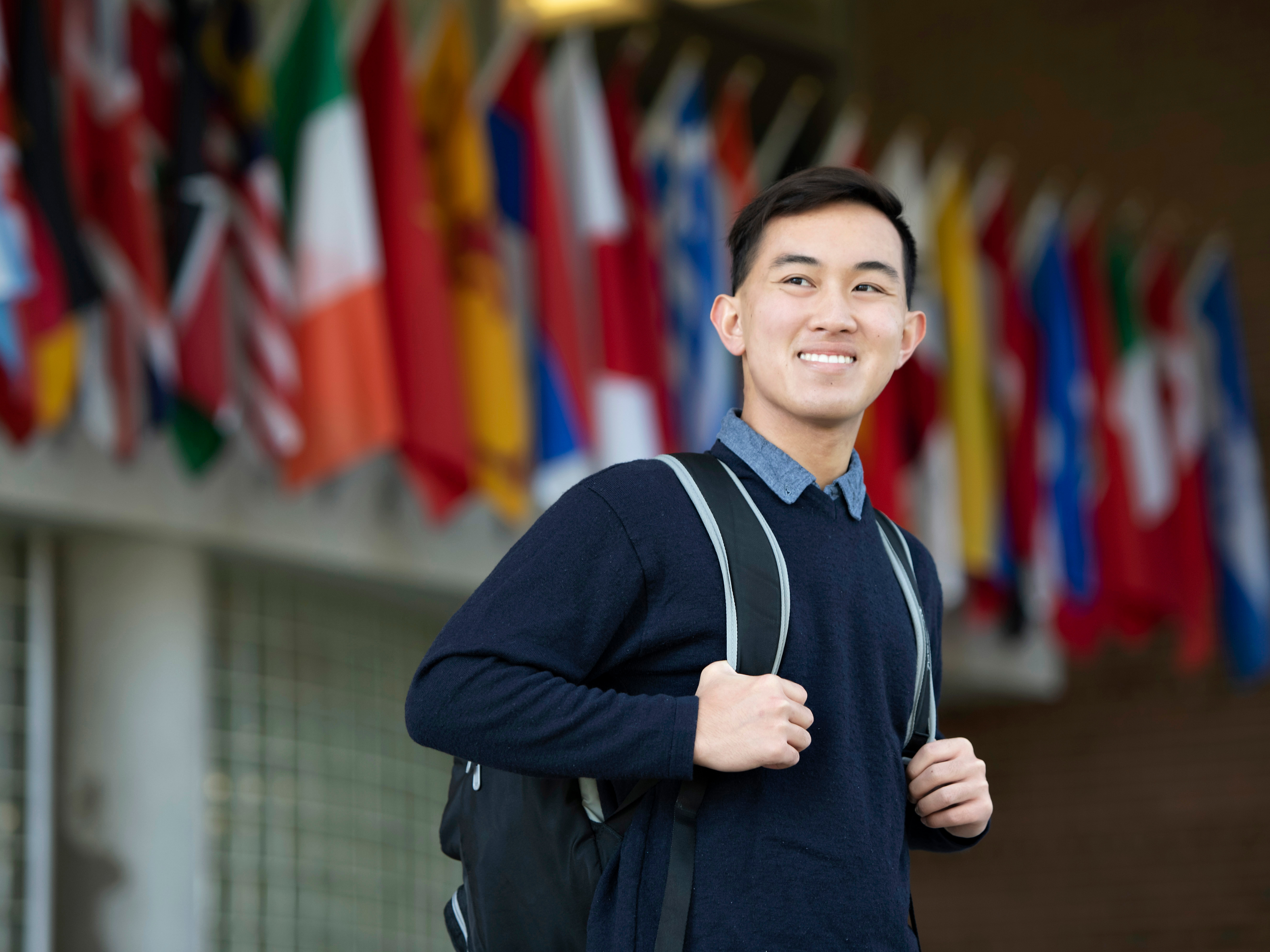 A Drake student with a backpack, hands looped around the straps, stands in front of a building with international flags on the exterior
