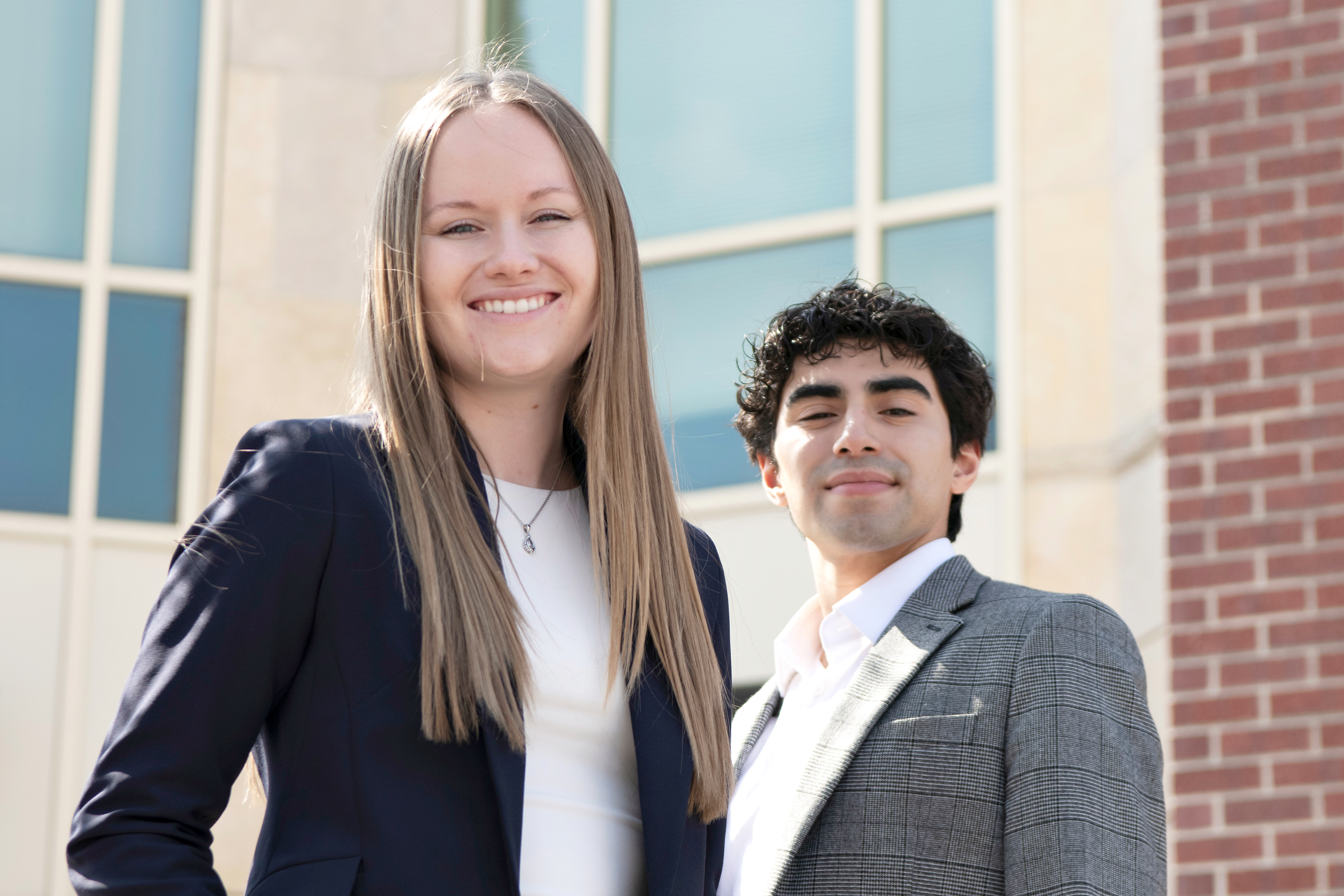Two Drake students in suits stand in front of a brick building with large windows