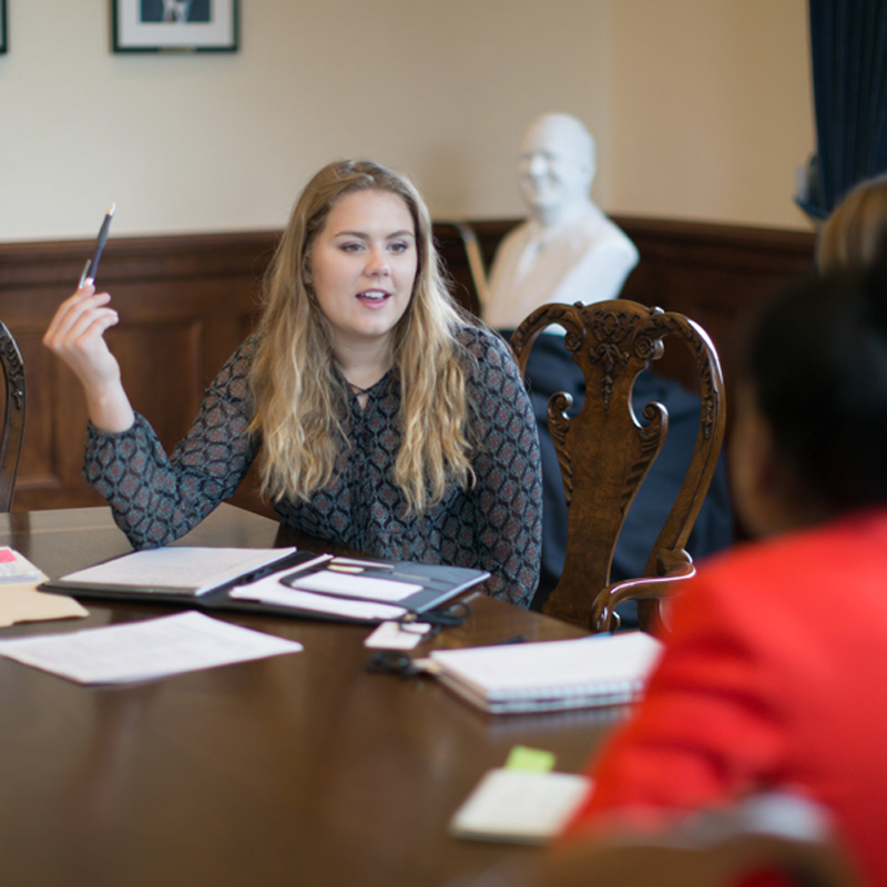 A student sits at a table, holding a pen and speaking to someone