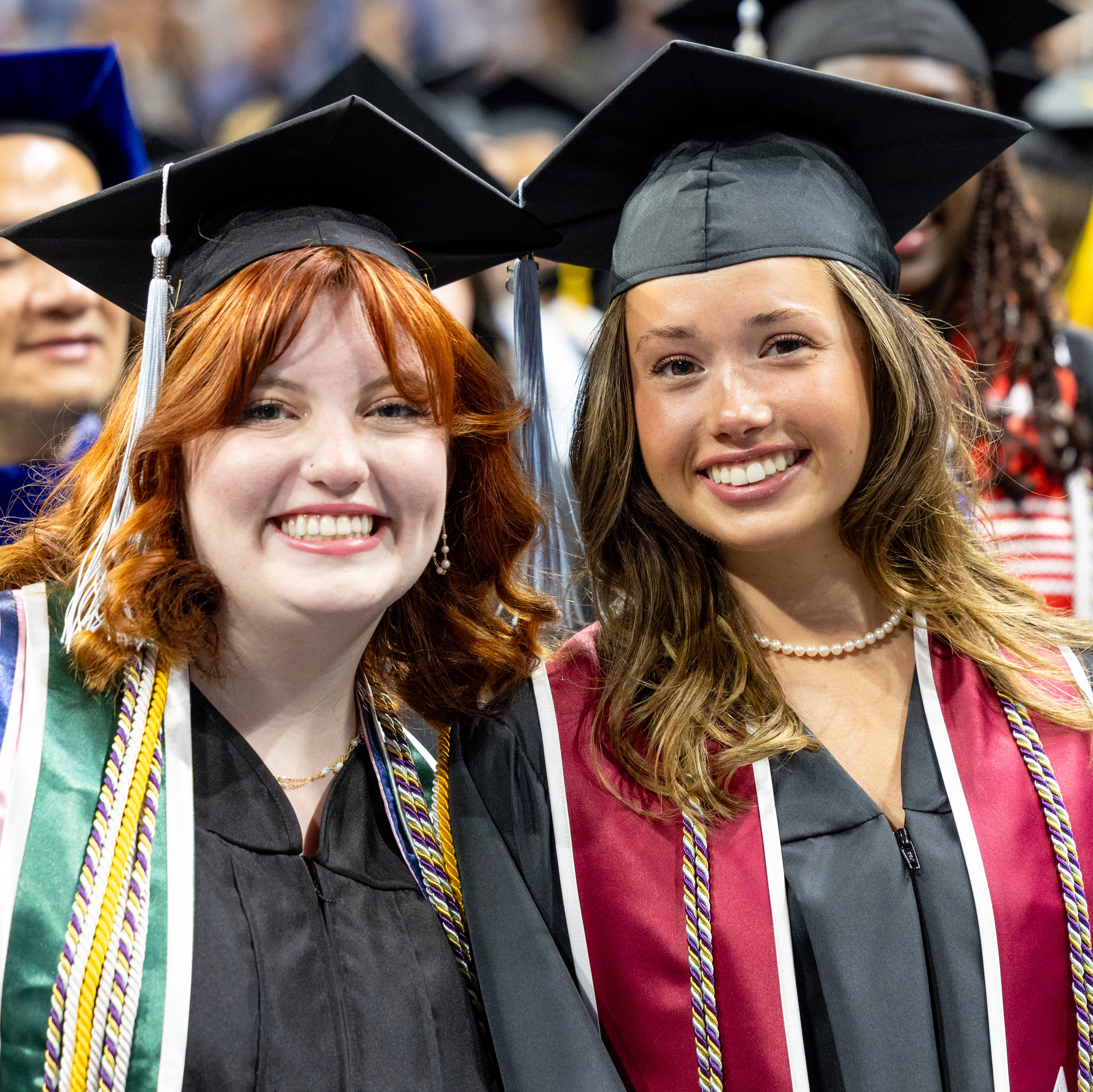 Two Drake students in caps, gowns, and honors sashes smile while standing close during commencement