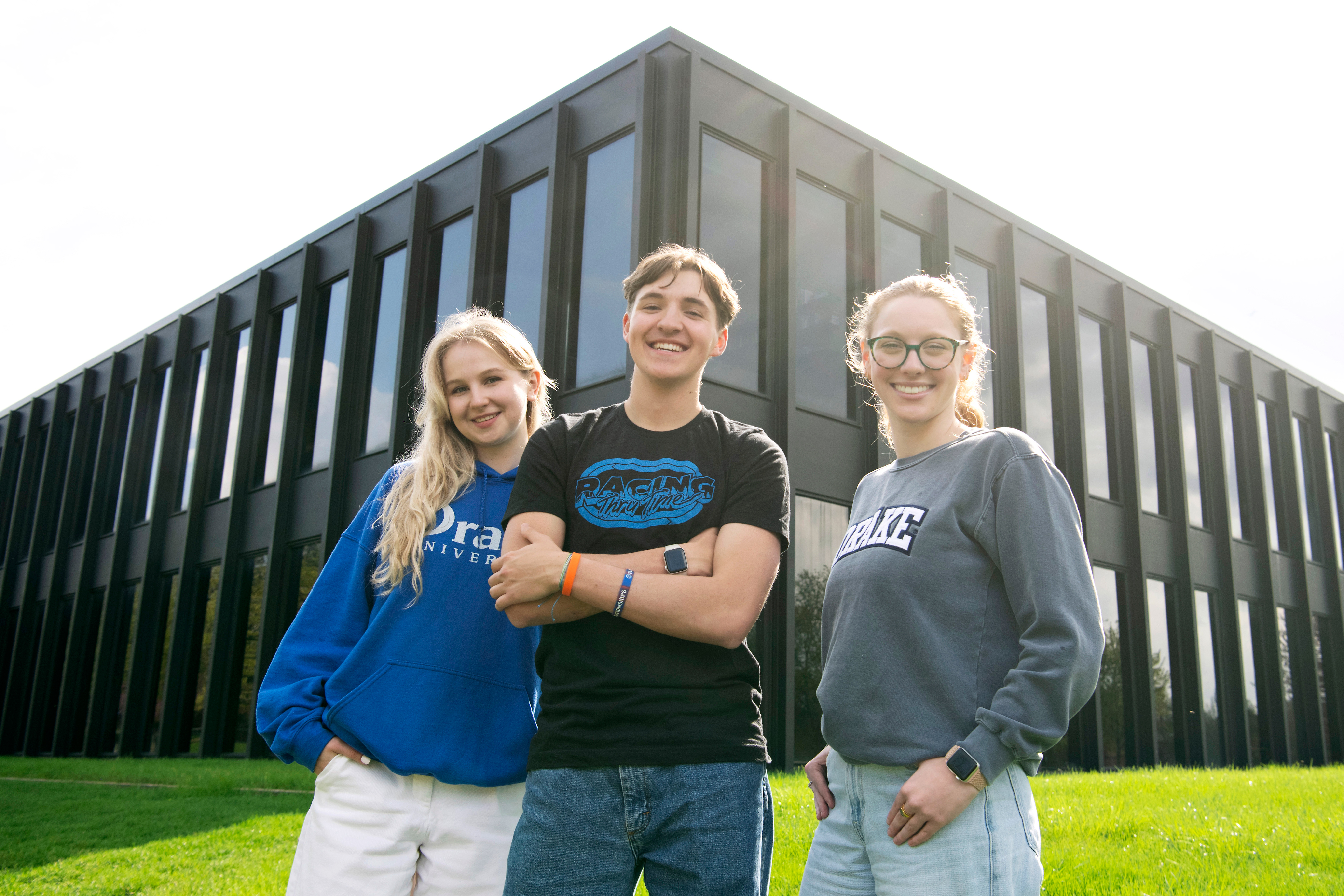 Three students stand on the grass by a corner of Drake's School of Journalism and Mass Communication building