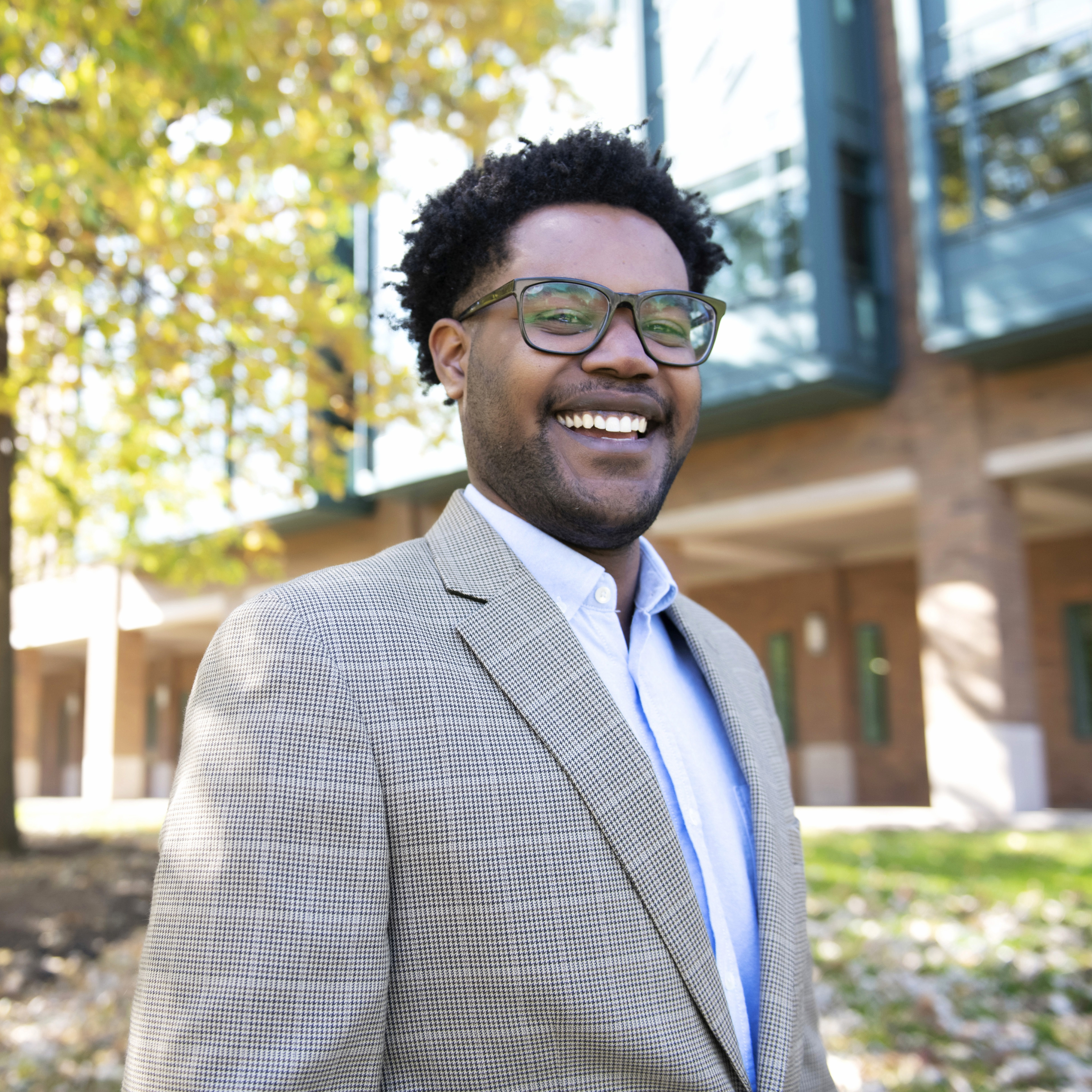 A Drake student in glasses wears a gray suit in front of a campus building on a sunny day