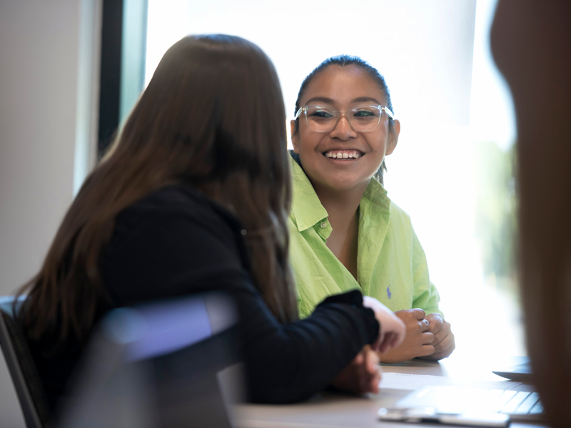 A student sits next to and smiles at another student who is facing away from the camera