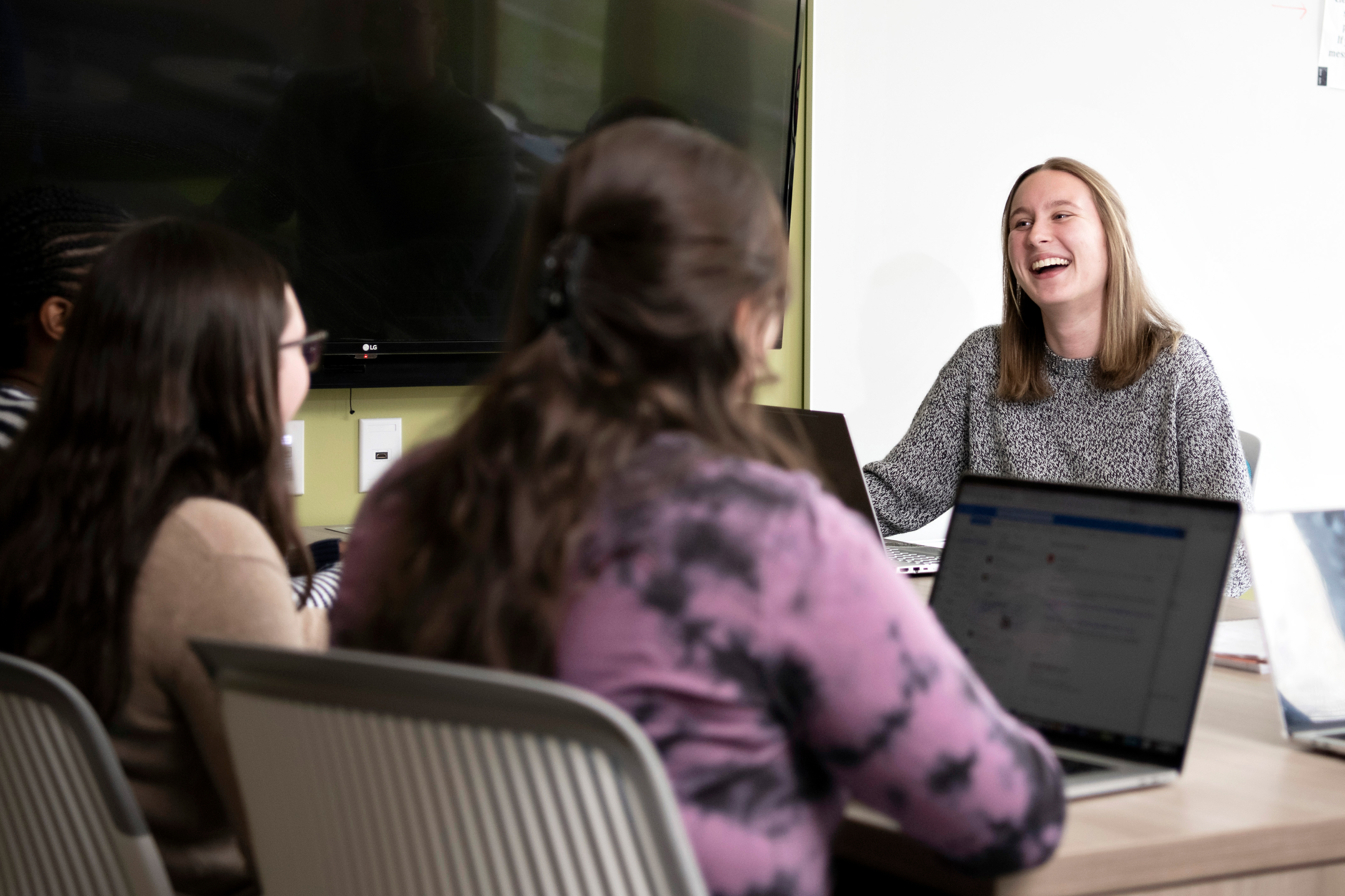 Students sit together at a table with laptops, one student is facing the camera while smiling and the others are facing away