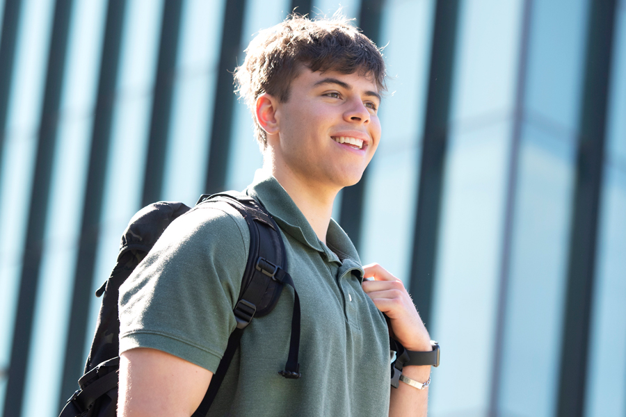 A student wearing a backpack looks off camera, smiling, while outside on campus