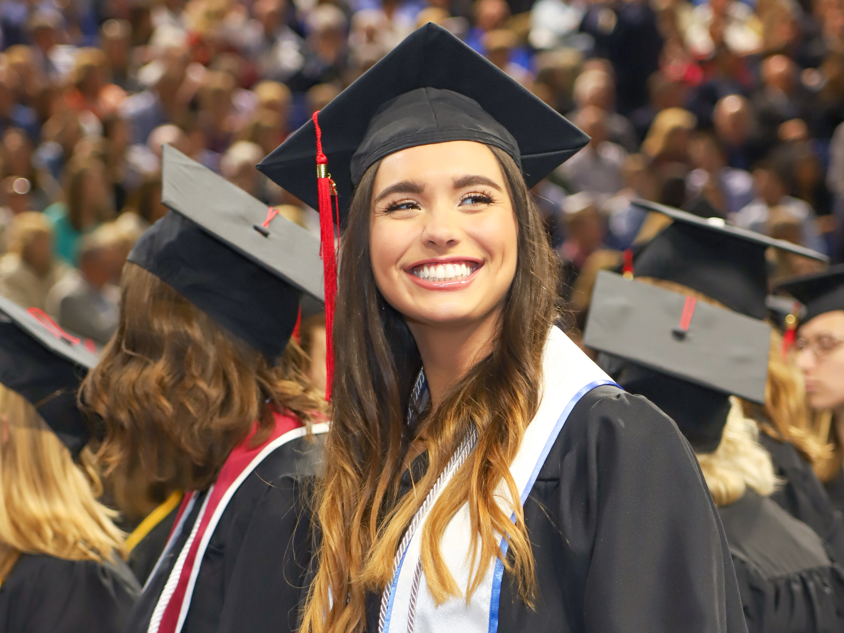 A Drake student in a cap, gown, and honors sash looks away from the crowd to smile during commencement