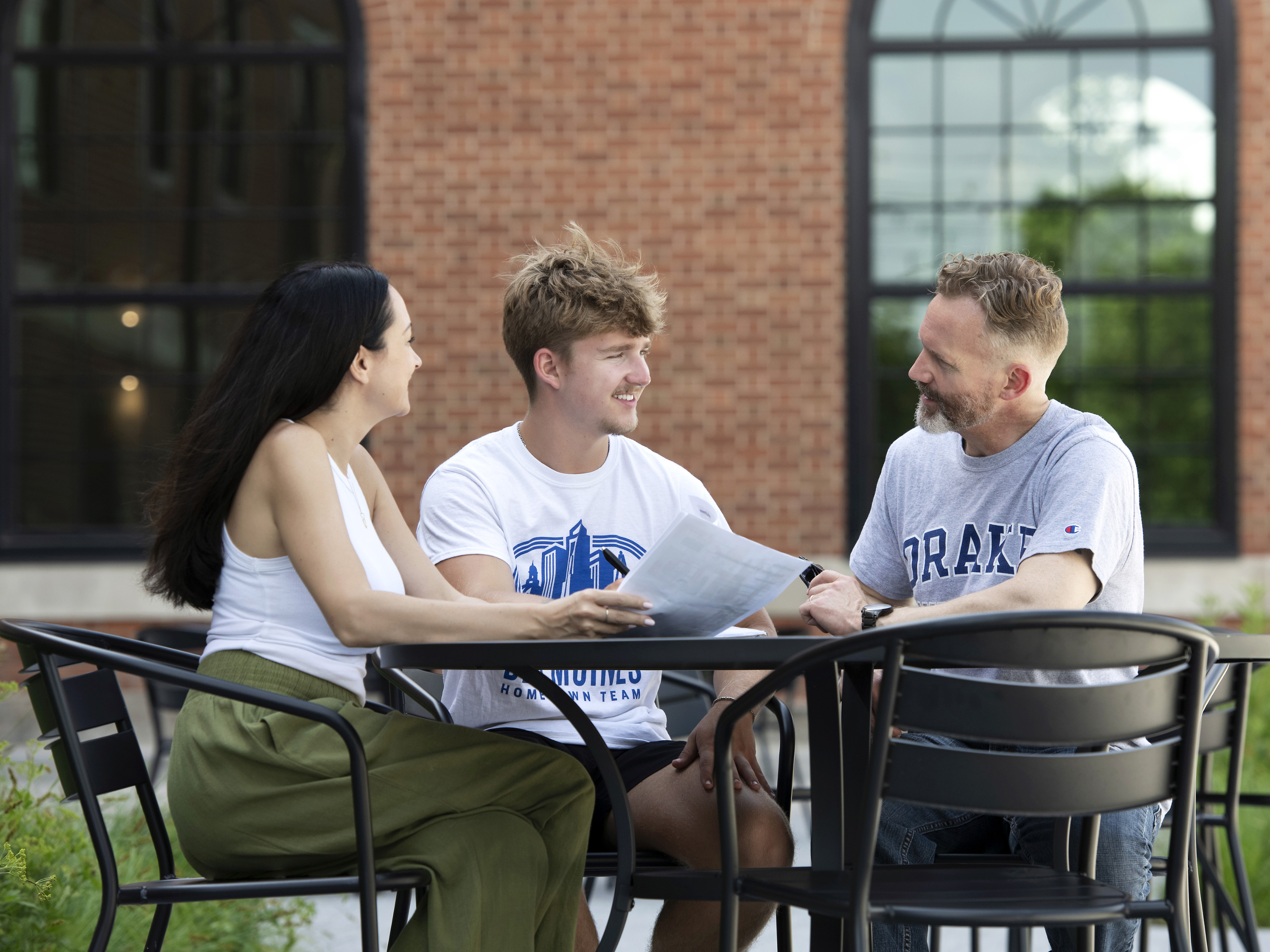 Two Drake students and a faculty member meet around a table outdoors to discuss an assignment