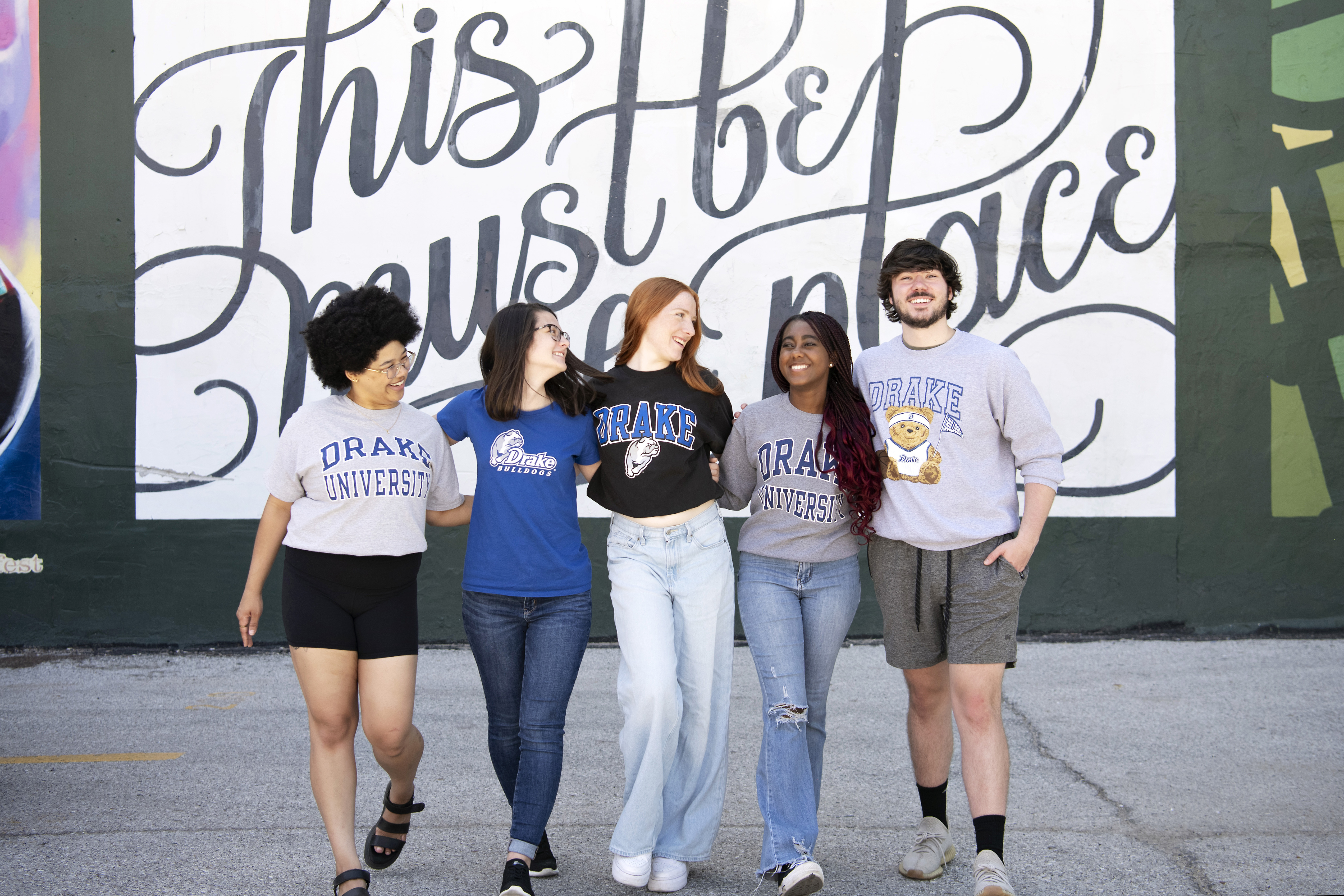A group of Drake students stand arm in arm in front of an outdoor mural that reads "This must be the place."