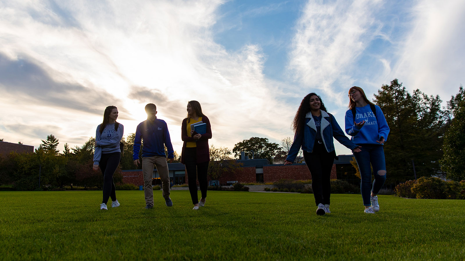 As the sun sets behind trees and classroom buildings, a group of Drake first-year students walk through a grass field