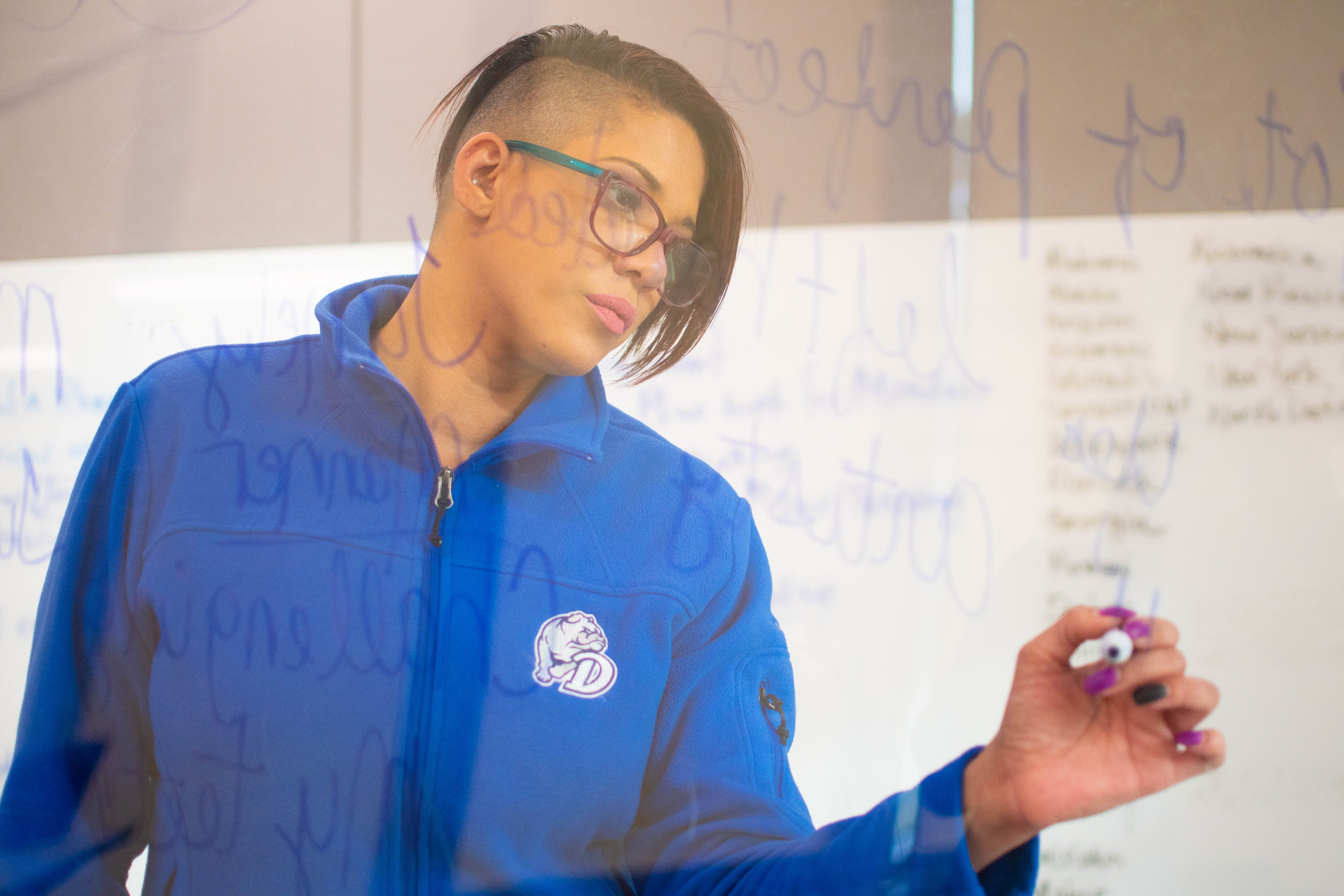 A student in glasses and a blue zip-up jacket scrawls notes on a glass brainstorming board in a Drake classroom