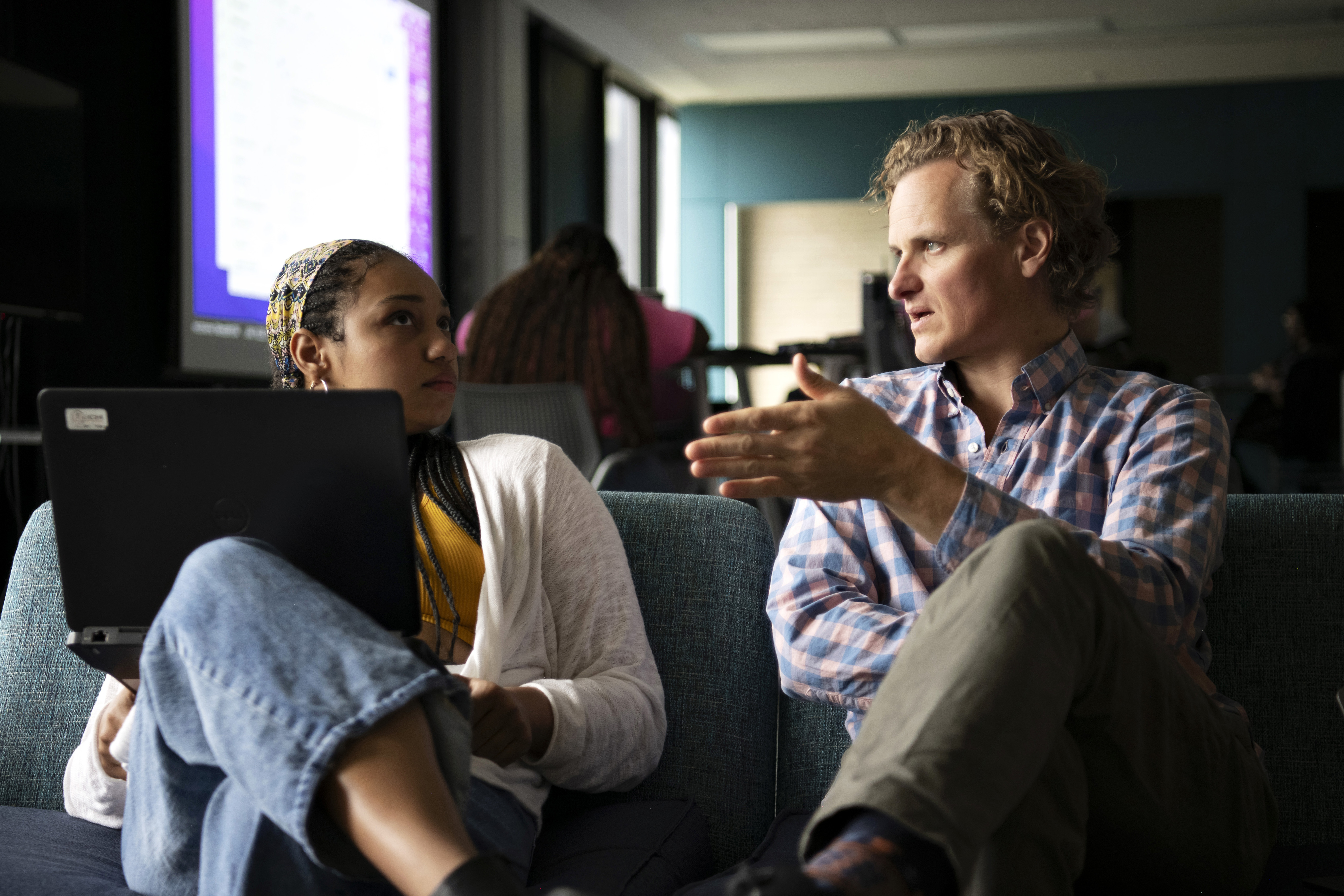 A faculty member holds a meeting with a Drake student, supporting a laptop, in a common area with couches