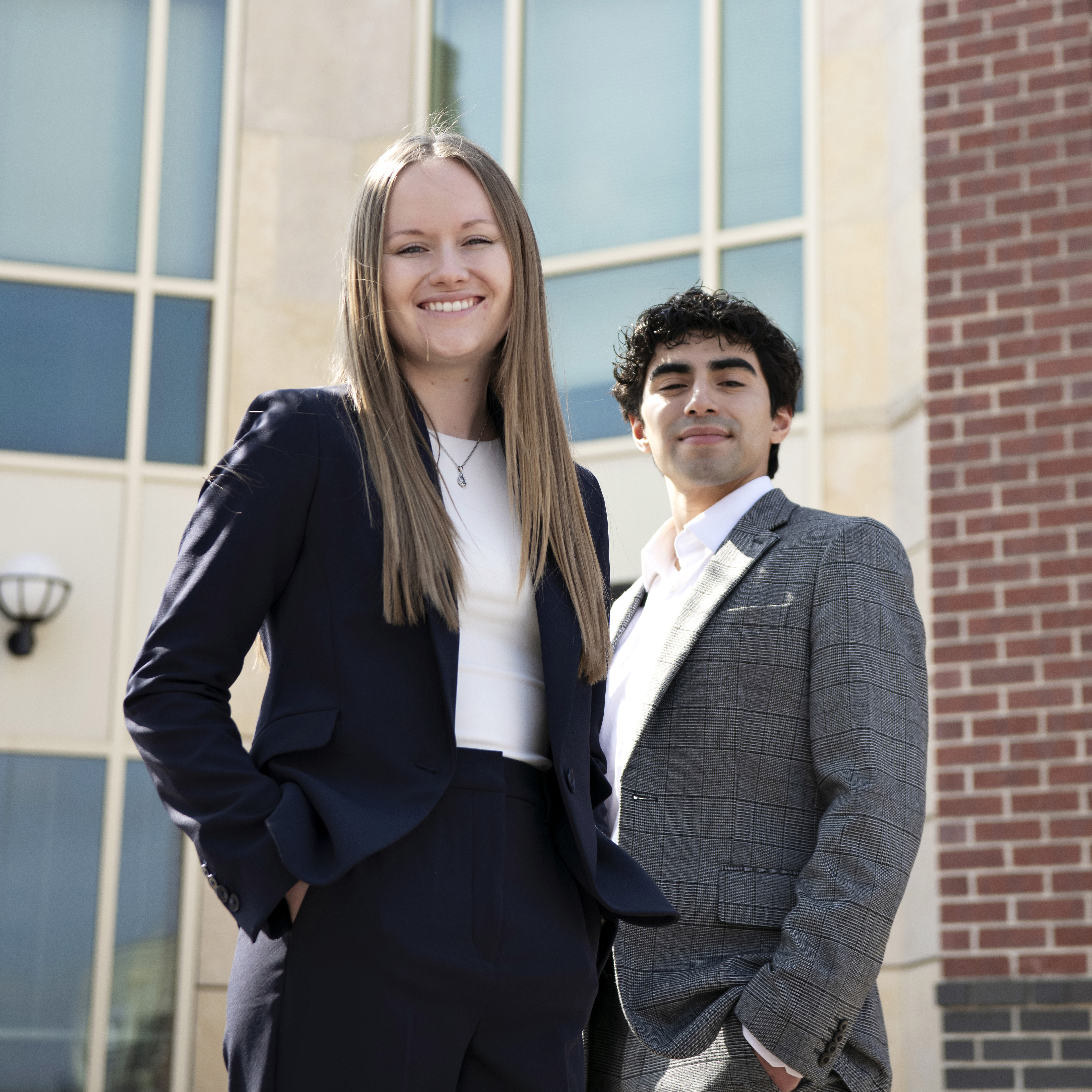 Two Drake Law students in blazers stand in front of the Law School building, also the home of the Drake Legal Clinic