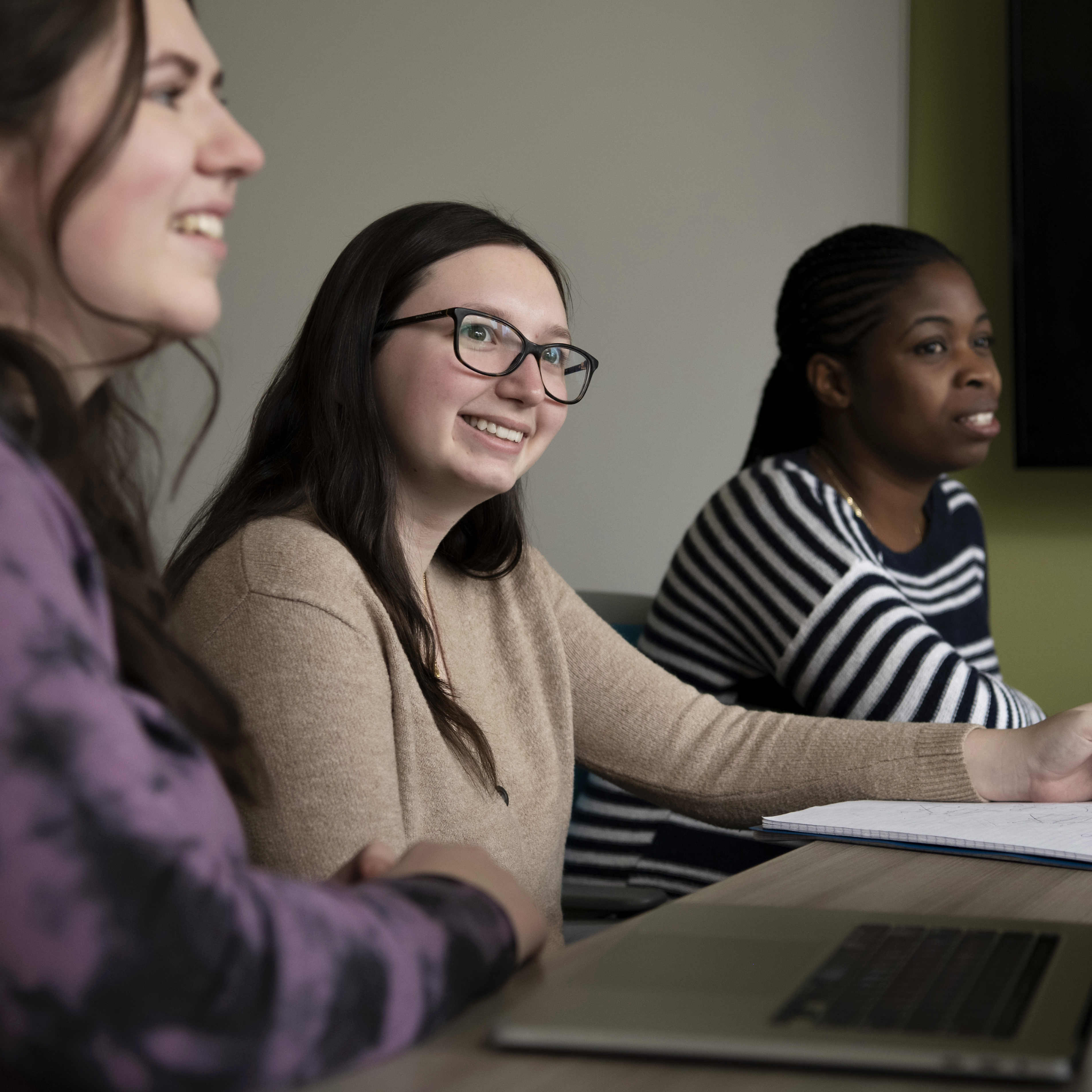 Graduate students, seated around a table with their laptops, smile and focus in a Drake classroom