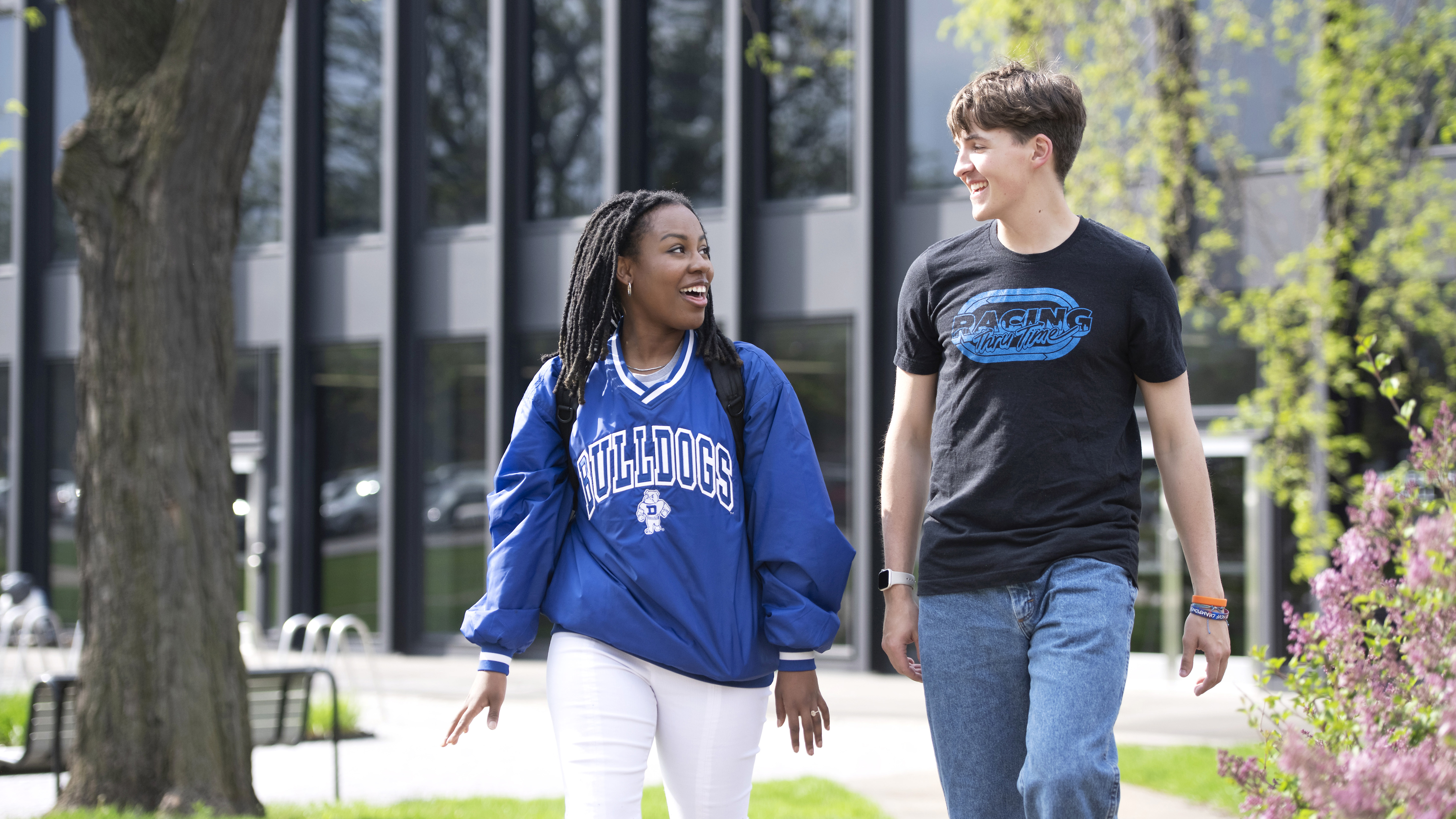 Two Drake international students, including one in a nylon school pullover, have a casual conversation while walking near a windowed classroom building