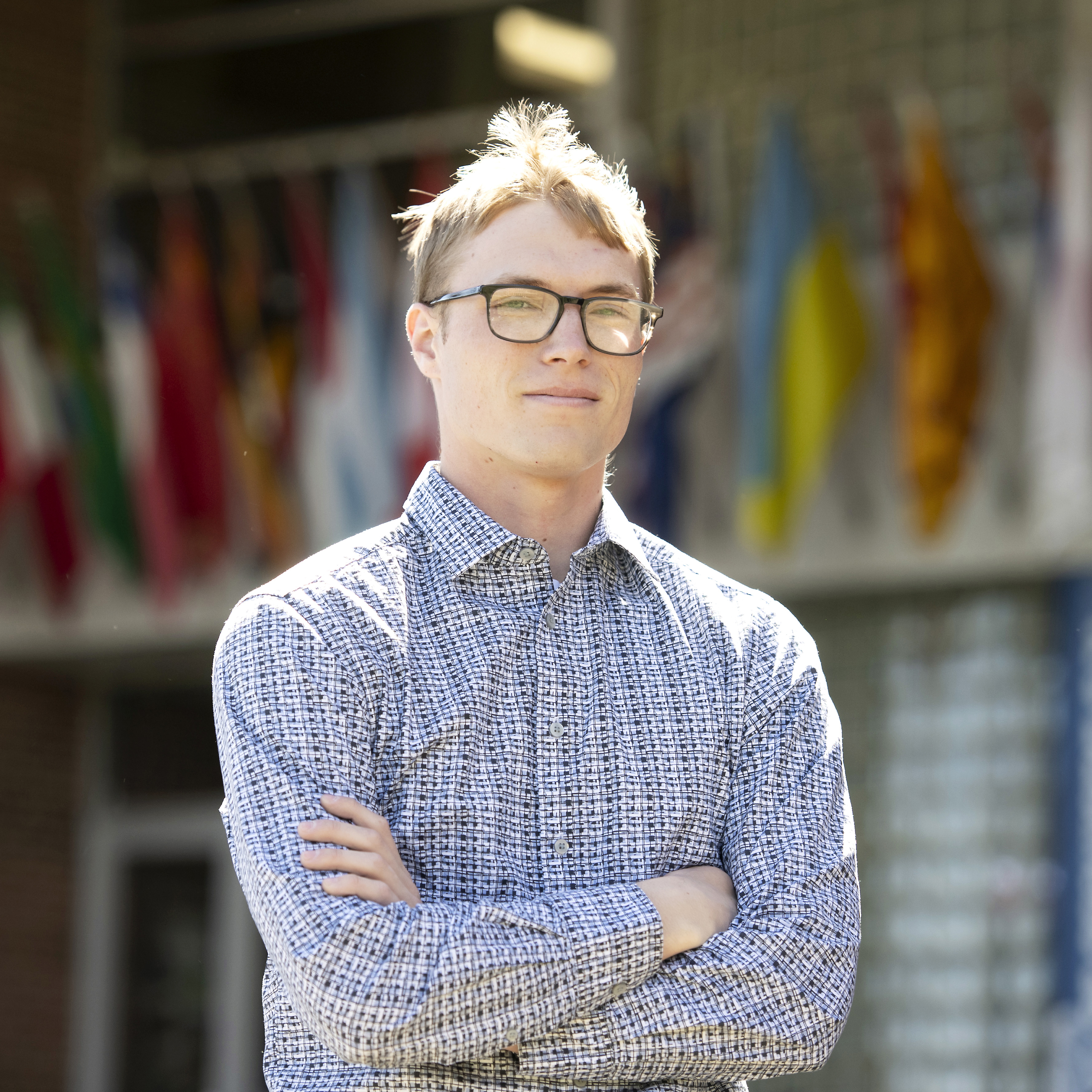 A Drake student in a patterned button-down and glasses stands in front of a building that features several international flags