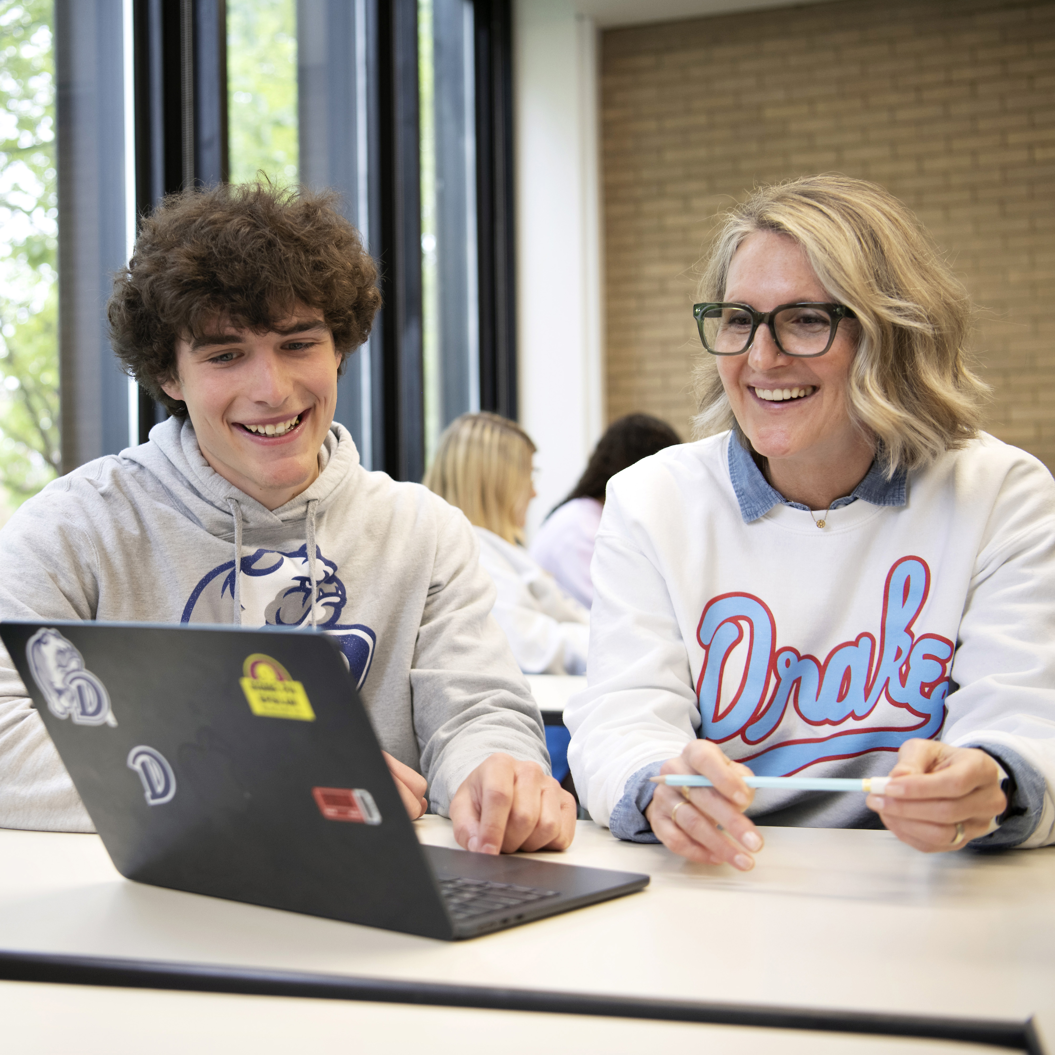 A professor helps a student better understand a concept, both wearing Drake sweatshirts while in front of a laptop