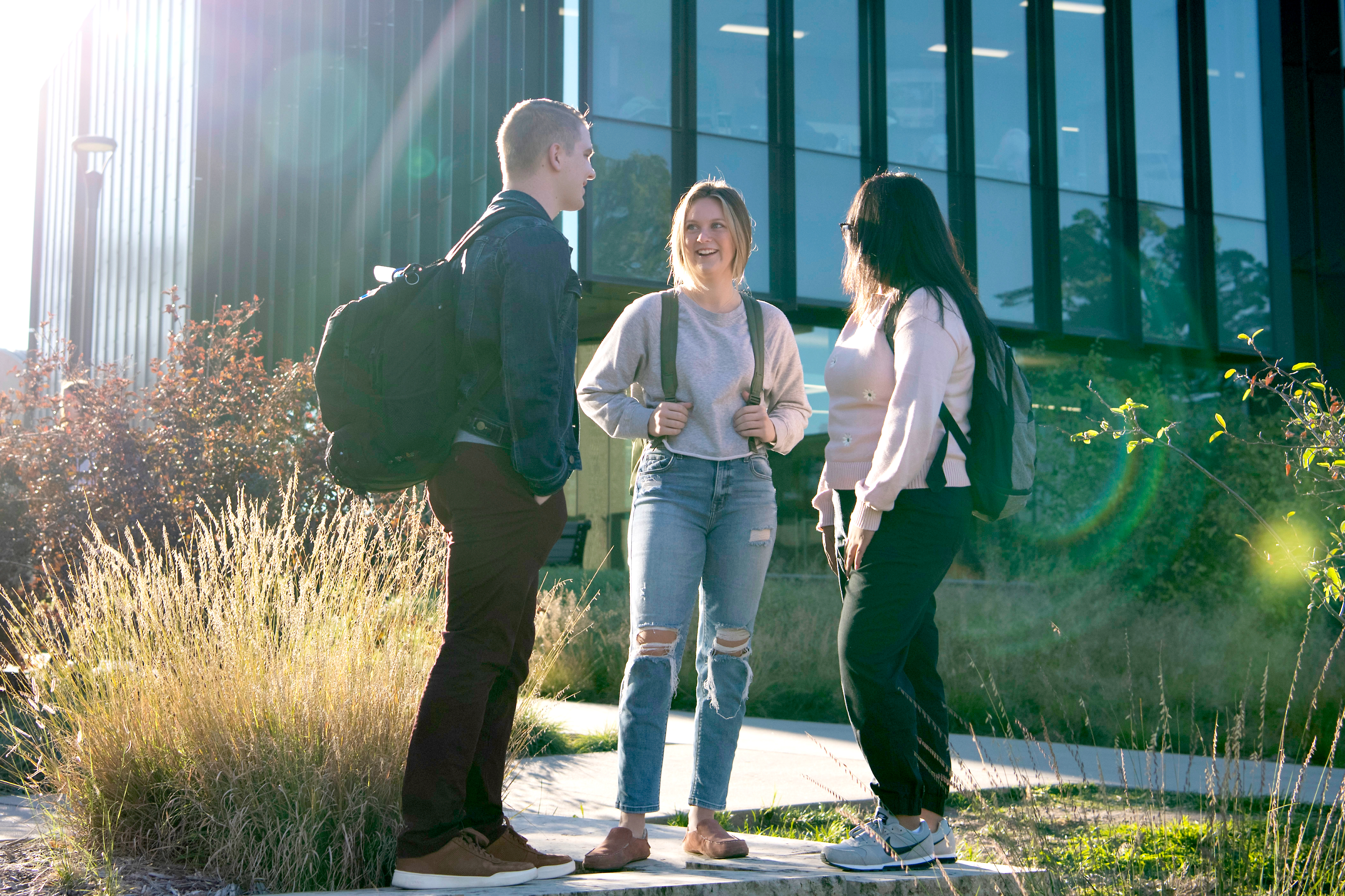 Three students wearing backpacks, stand while conversing outside a building on campus