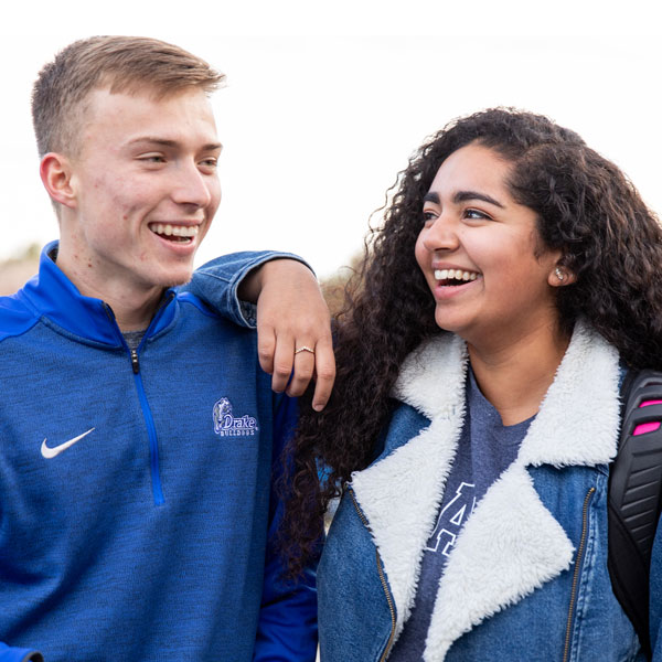 Two students smiling and talking outdoors, one with an arm around the other’s shoulder.