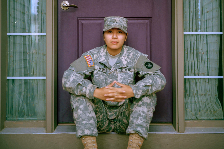 A student in U.S. Army camoflauge attire sits on a doorstep while looking into the camera
