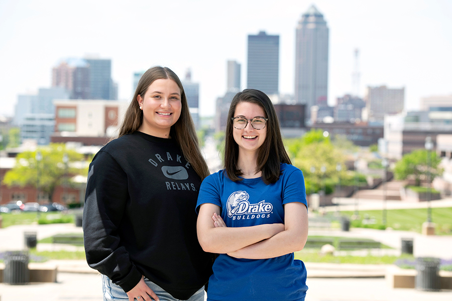 Two recent Drake alumni in school shirts stand, one with their arms crossed, near the Des Moines skyline