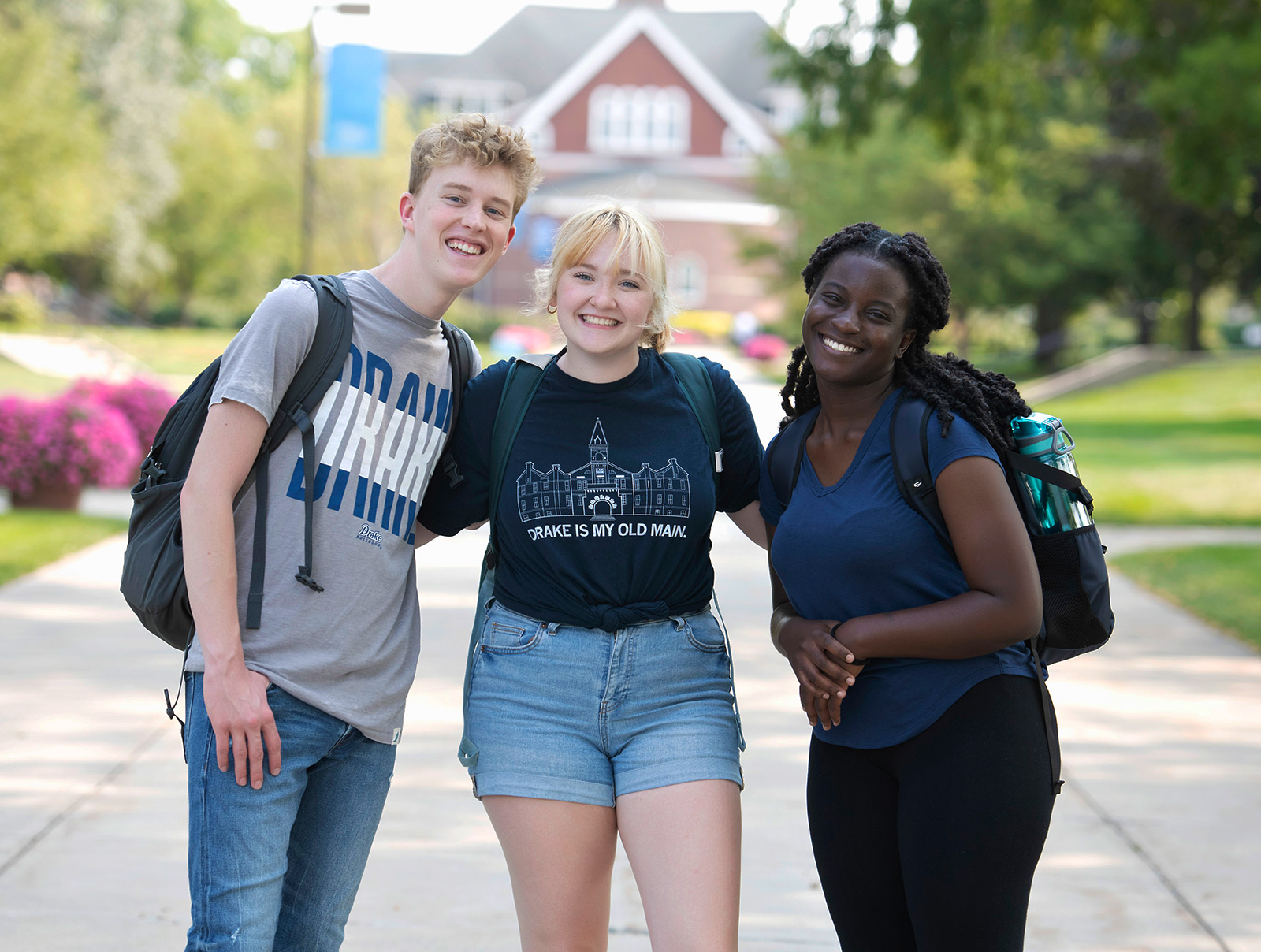 Three drake students standing together smiling on the sidewalk between Harmon Fine Arts Center and Opperman Hall with Sheslow in the background.