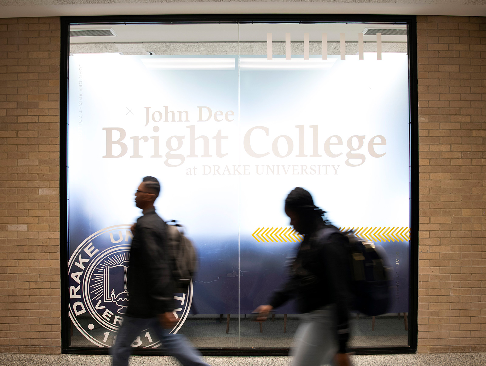 Two students walking down a hall with the John Dee Bright College logo on a window behind them.