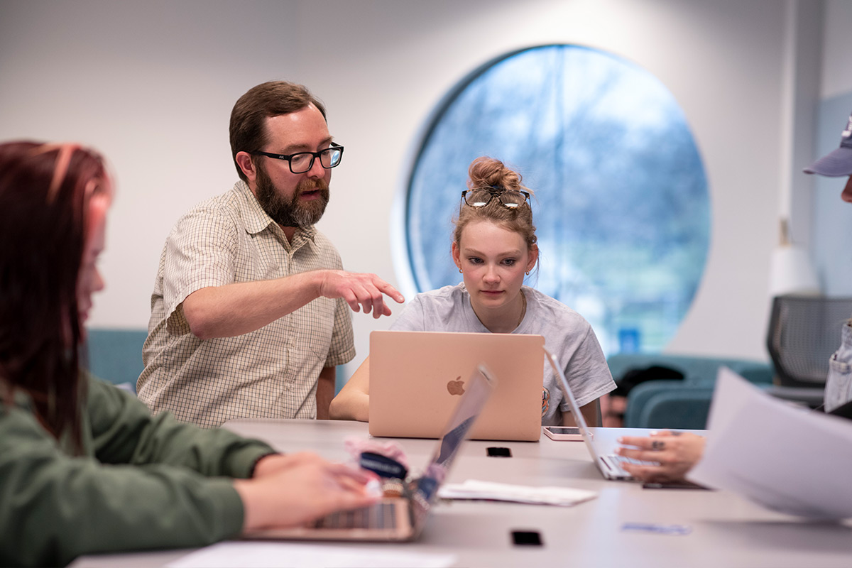 Drake University faculty points to a laptop over a student's shoulder while sitting at a table.
