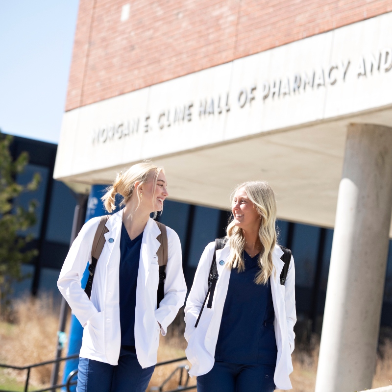 Two Drake students in lab coats converse outside the College of Pharmacy and Health Sciences building