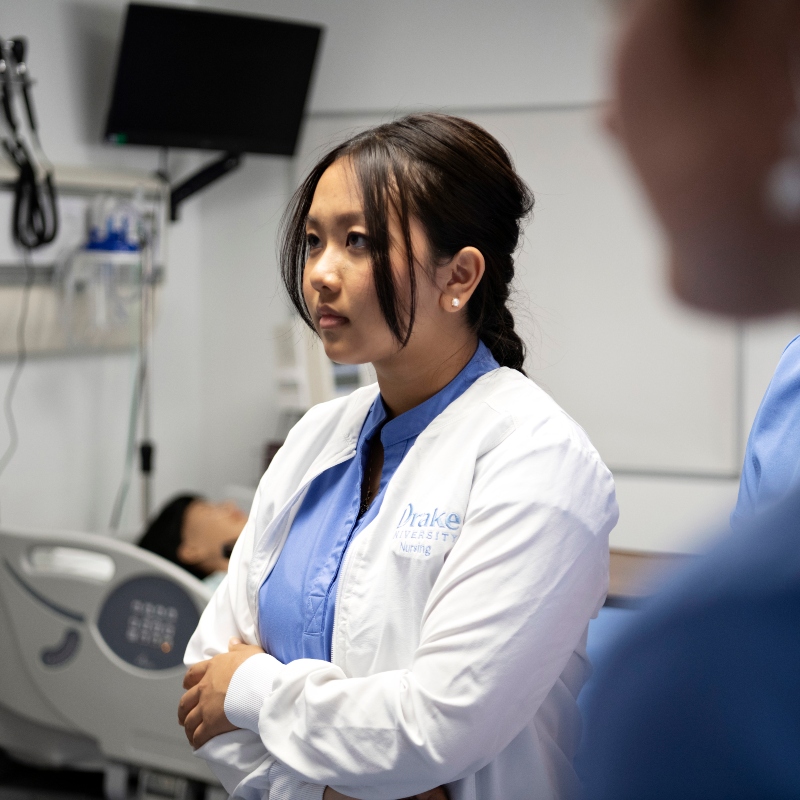 A Drake student in a lab coat folds their arms while training in one of the College of Pharmacy and Health Sciences' simulation spaces
