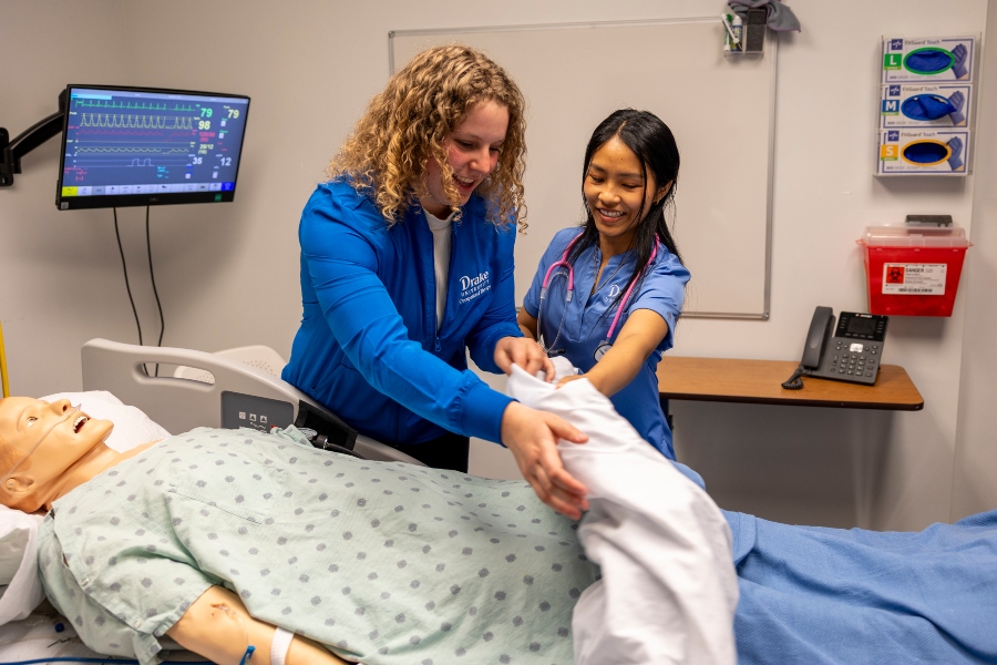 An instructor and a Drake student with a stethoscope lift a blanket off a manikin in one of the College of Pharmacy and Health Sciences' simulation labs