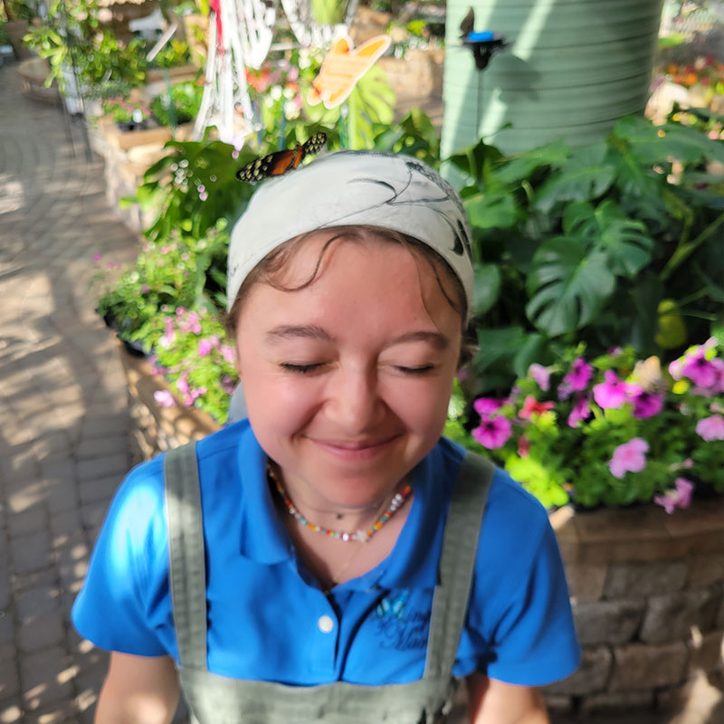 Allie Raines with eyes closed as a butterfly rests on her headband in a lush garden setting.