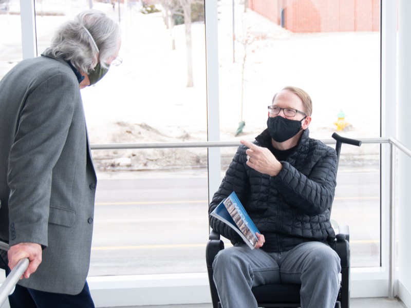 A man stands speaking to another man who is seated in a wheelchair and carrying a book