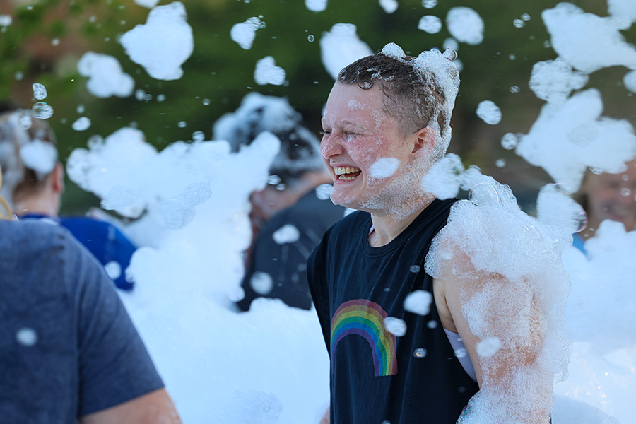 A Drake student in a rainbow T-shirt smiles as they emerge from a cloud of foam during a campus event