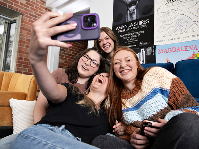 Four Drake students take a selfie with a smartphone in a campus common area with couches and posters on the walls