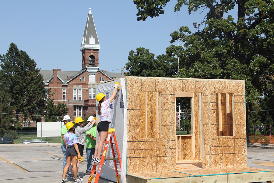 Standing outdoors, Drake University student volunteers in hard hats apply a layer of Tyvek to a plywood structural frame