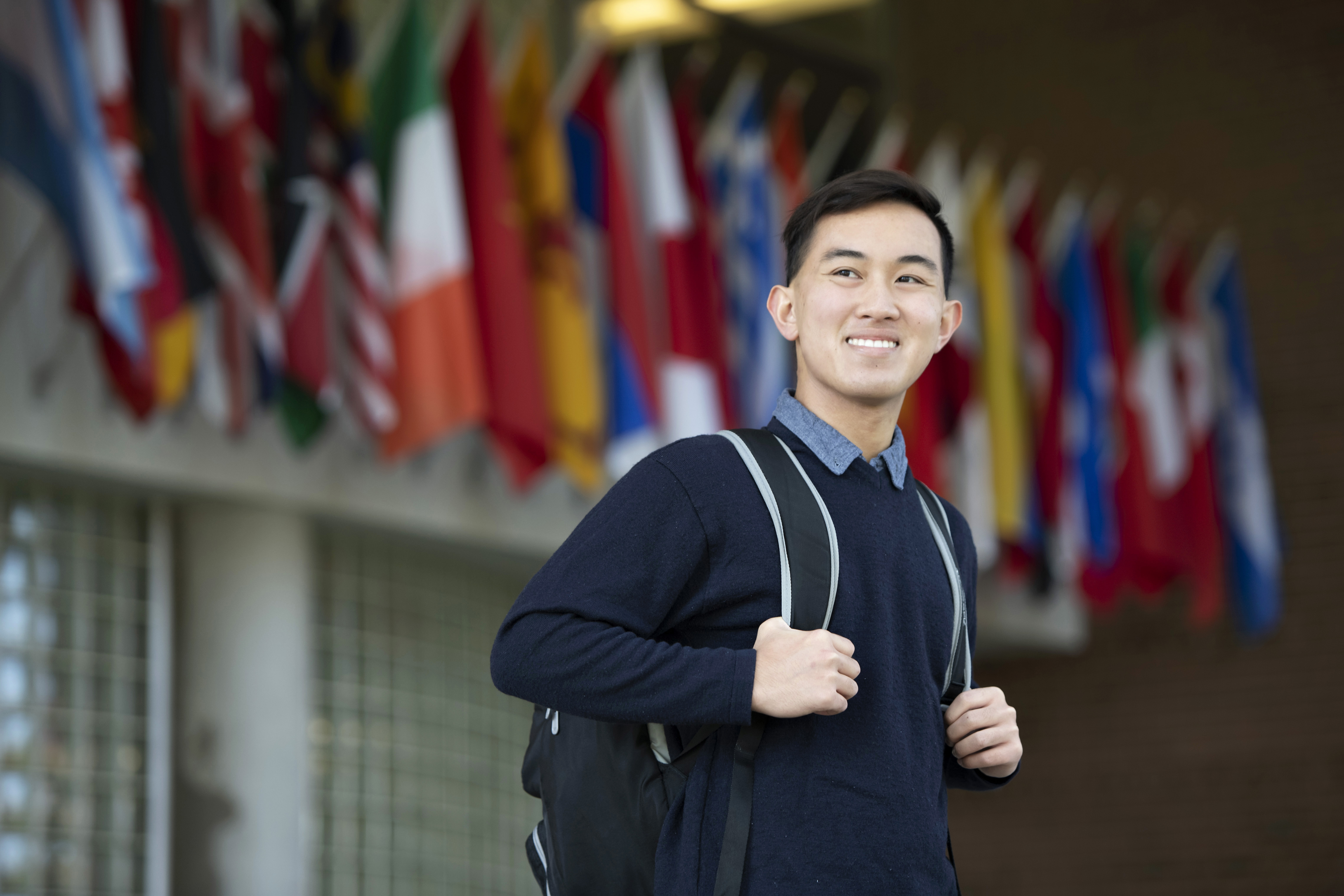 A Drake student, with their thumbs hooked around their backpack straps, stands in front of a series of international flags outdoors