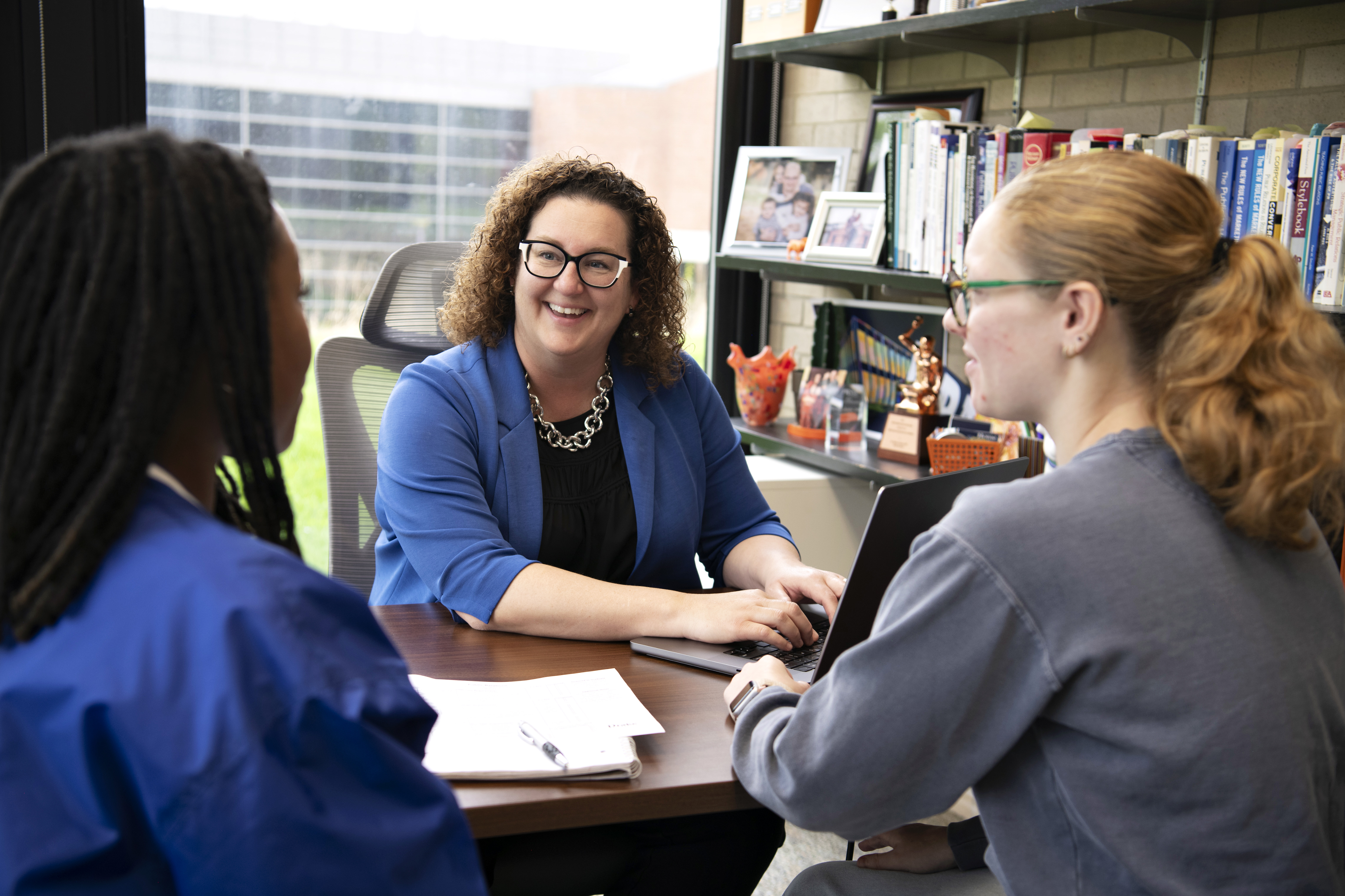 An advisor, sitting behind a desk and typing on a laptop, meets with two Drake students