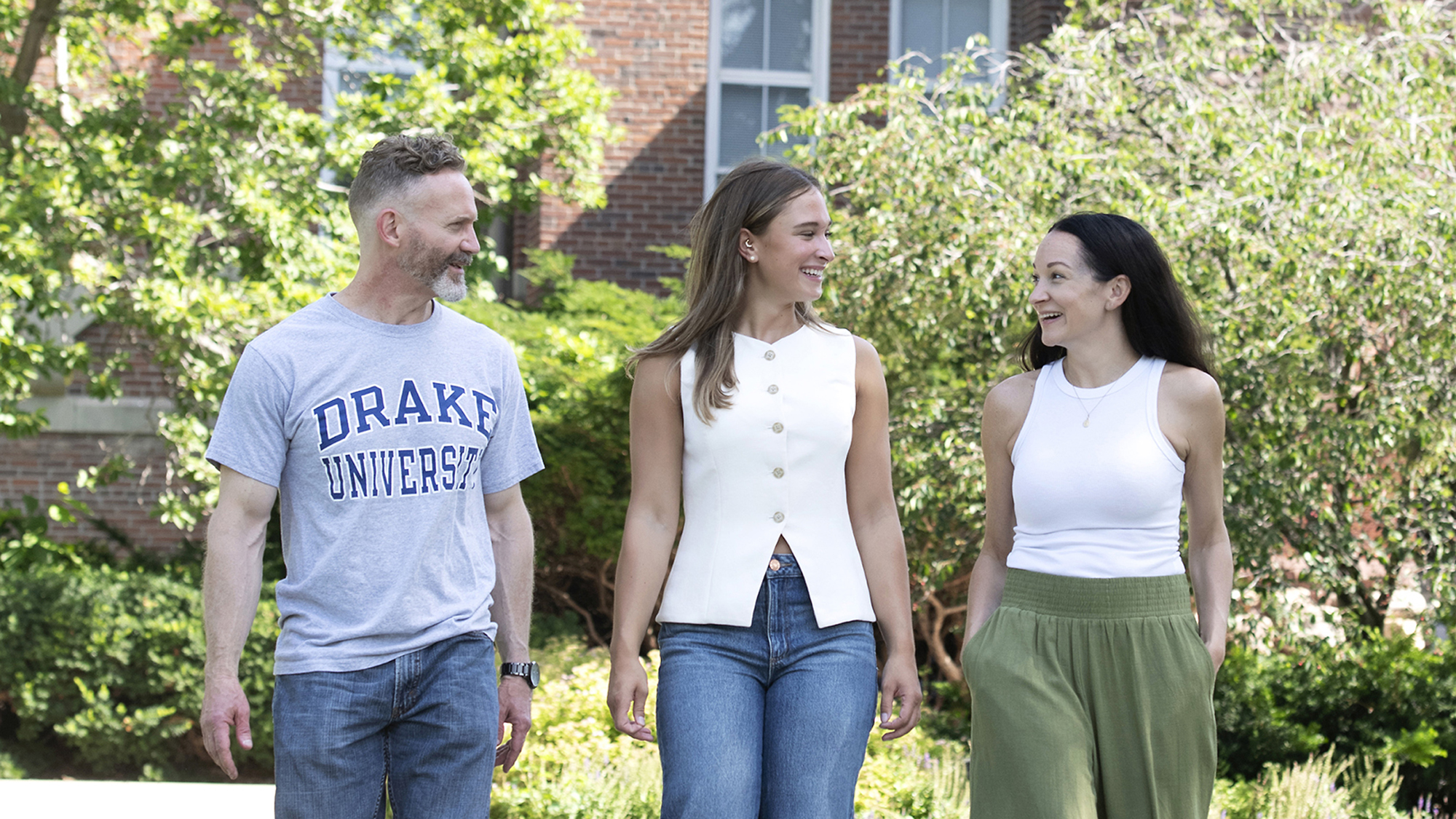 Standing near a brick building and bushes, a parent and their student enjoy a personal tour by a current Drake student