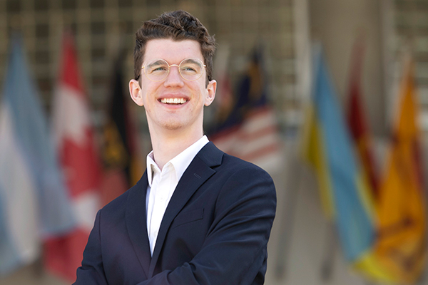 A Drake Business/Law 3+3 student in glasses and a blazer crosses their arms and looks to the right while standing in front of international flags