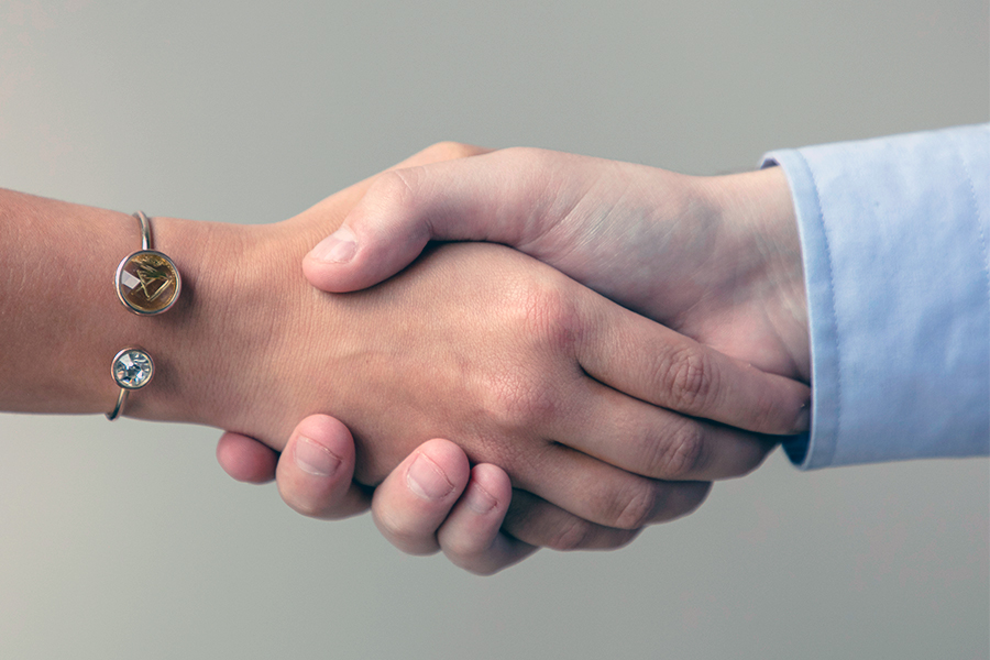 Two hands, one with a jeweled bracelet and another with a light blue shirt cuff, engage in a handshake
