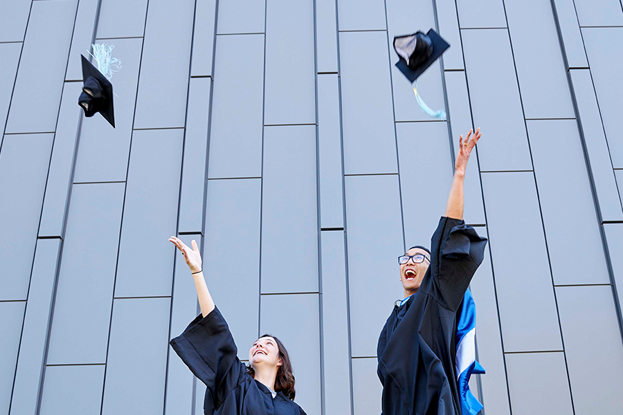 Two Drake students in graduation gowns throw their caps in the air in front of a building with an unevenly paneled exterior