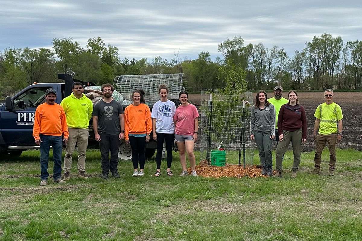 Group of 10 people standing around a tree recently planted in the new Perry Fruit Orchard.