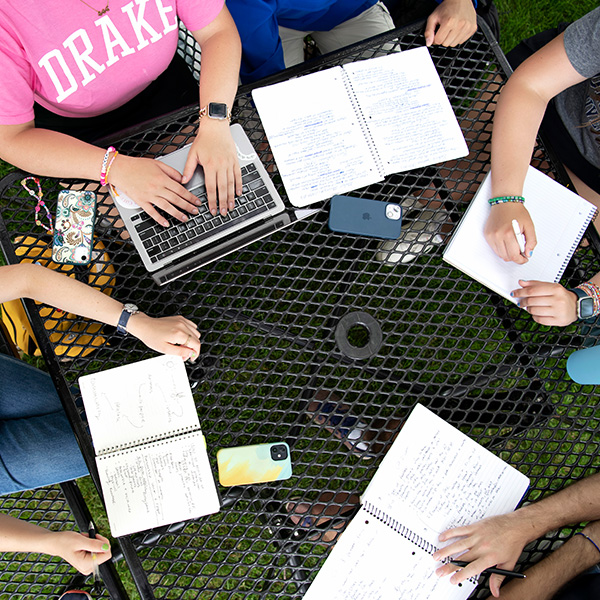 Aerial view above a table outdoors with students working together both on laptops and in notebooks.