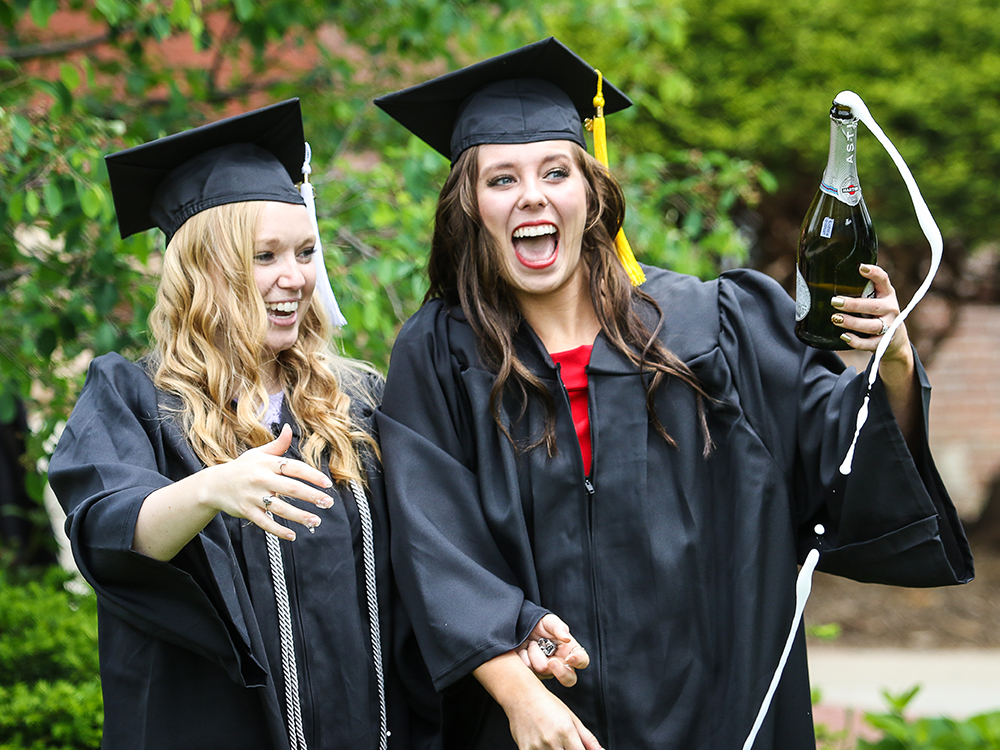 Two girls smiling in their regalia after commencement while holding a bottle of champagne in front of Old Main.