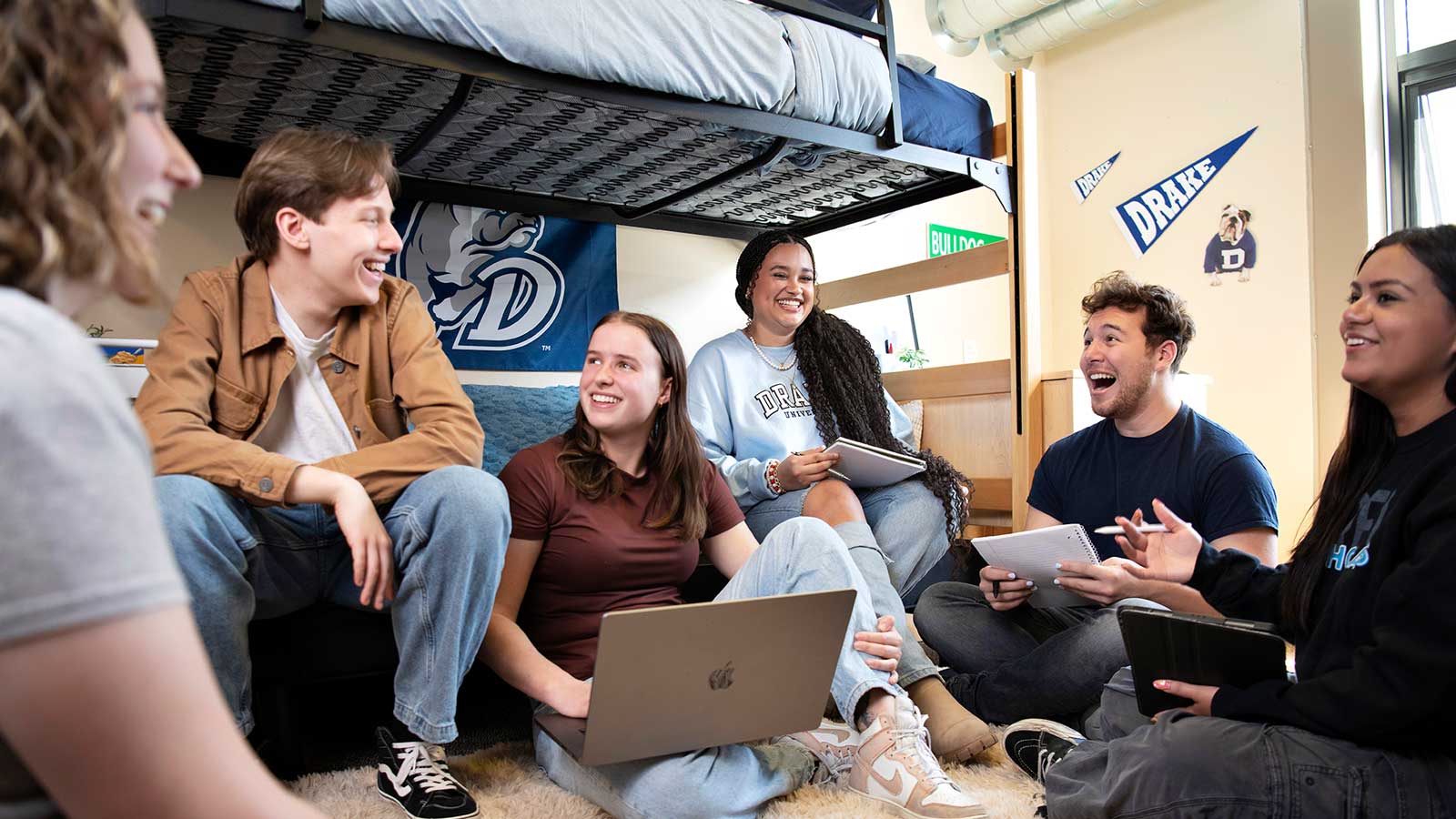 Six Drake University students sitting around on the ground of a dorm room laughing together.